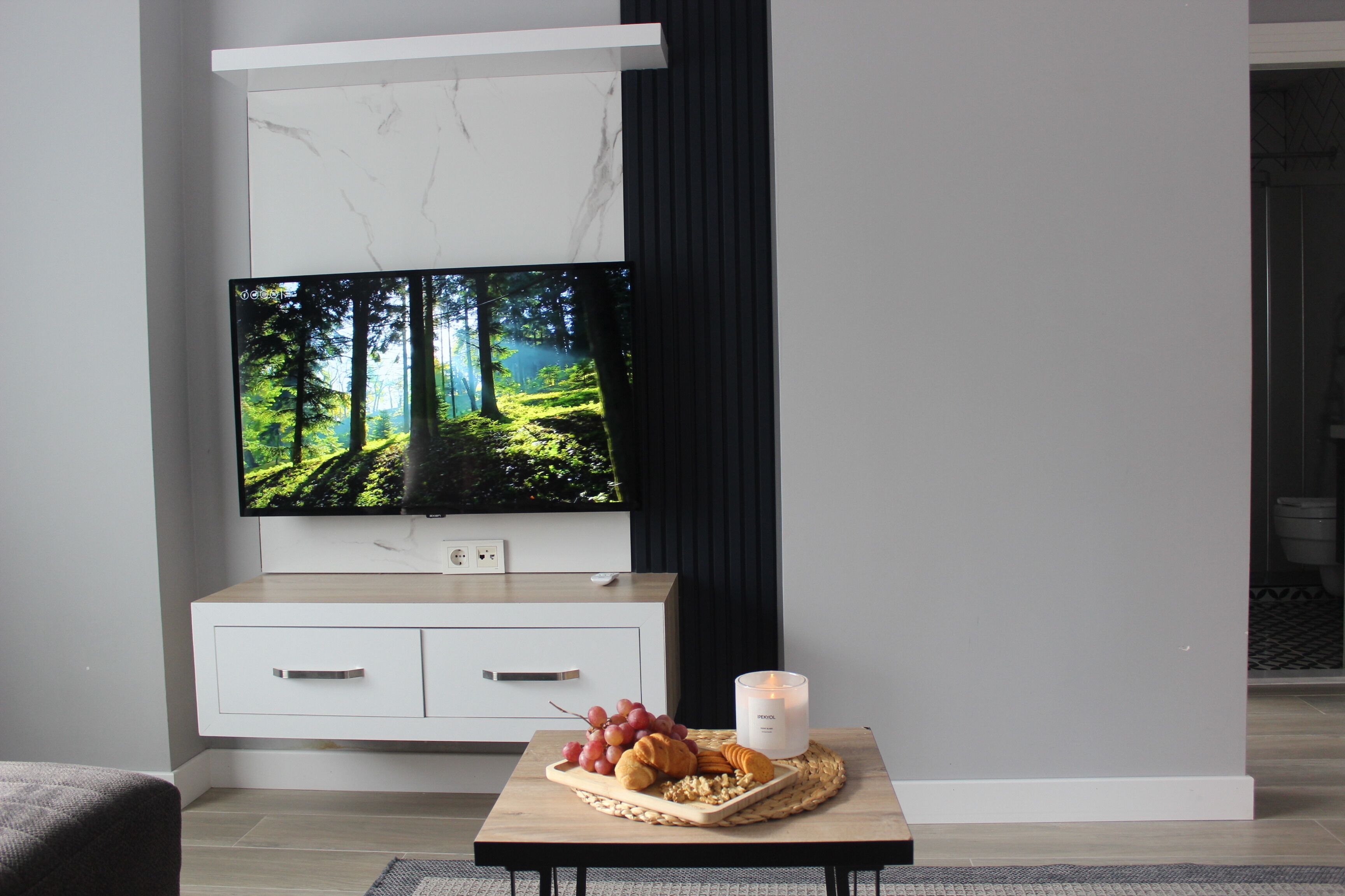 Un salon moderne avec un téléviseur à écran plat fixé au mur, une table basse en bois sur laquelle sont disposés des en-cas, et un meuble blanc avec des tiroirs.