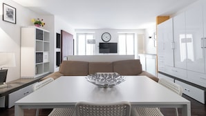 Una sala moderna con una mesa de comedor blanca, un sofá beige y un cuenco decorativo como centro de mesa. Un televisor de pantalla plana está montado en la pared sobre el sofá.