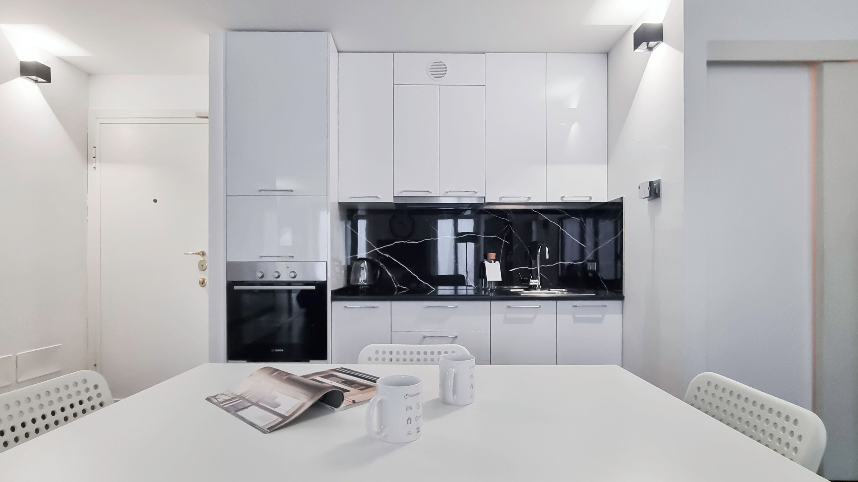 Une cuisine moderne avec des armoires blanches, un comptoir noir et un four encastré. On y trouve une table à manger avec deux tasses blanches et un magazine.