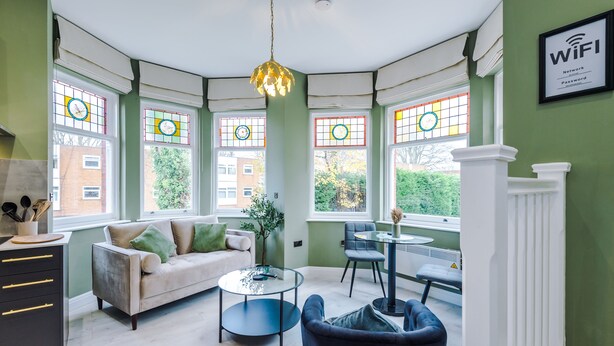 A modern living room with a sofa, a coffee table, and a chair. There are large windows with stained glass, a hanging light fixture, and a WiFi sign on the wall. - Hilltop Serviced Apartments - Stockport (Stockport)