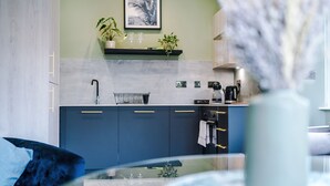 A modern kitchen with dark blue cabinets, a white countertop, and a green wall. There are two potted plants on a shelf above the counter. - Hilltop Serviced Apartments - Stockport (Stockport)