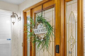 Une porte en bois avec une plaque indiquant « Maison de plage », une porte en verre ornée d’un motif décoratif et un luminaire fixé au mur.
