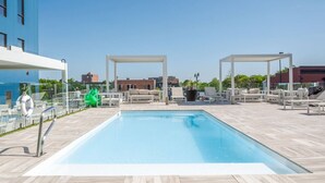 Piscina en la azotea con sillones de descanso y sombrillas, rodeada de edificios.