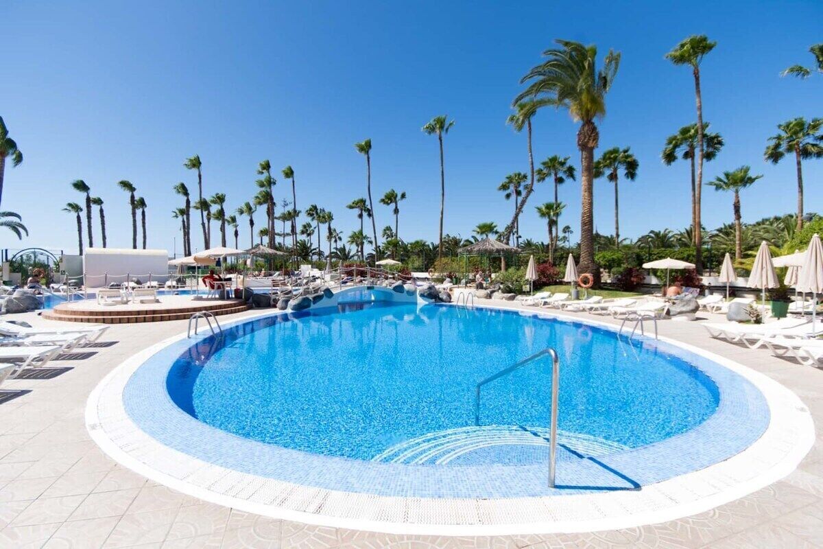 A circular pool with a clear blue center, surrounded by lounge chairs and umbrellas, with palm trees in the background.