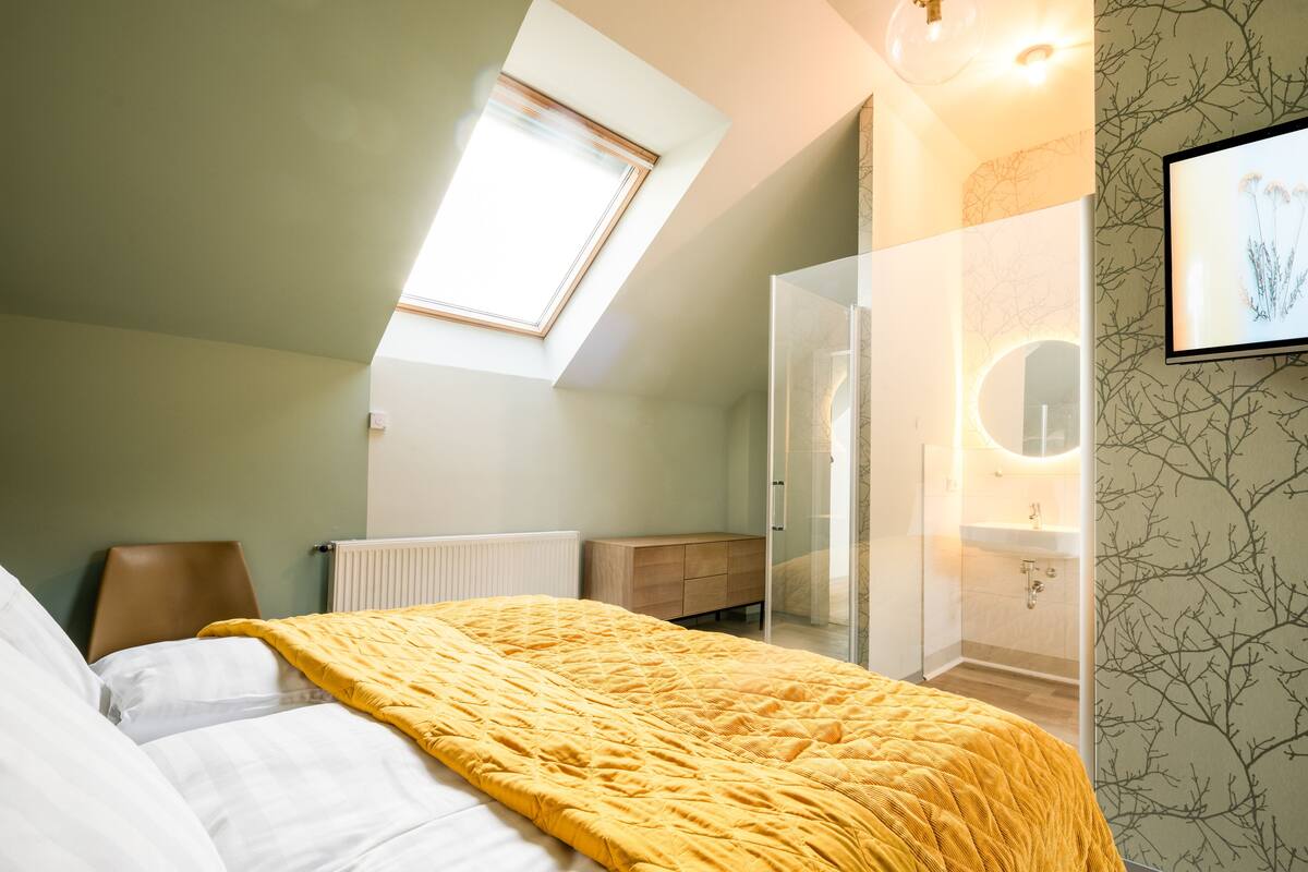 A modern bedroom with a skylight, a bed with a yellow blanket, a wooden nightstand, and a bathroom with a mirror and sink.