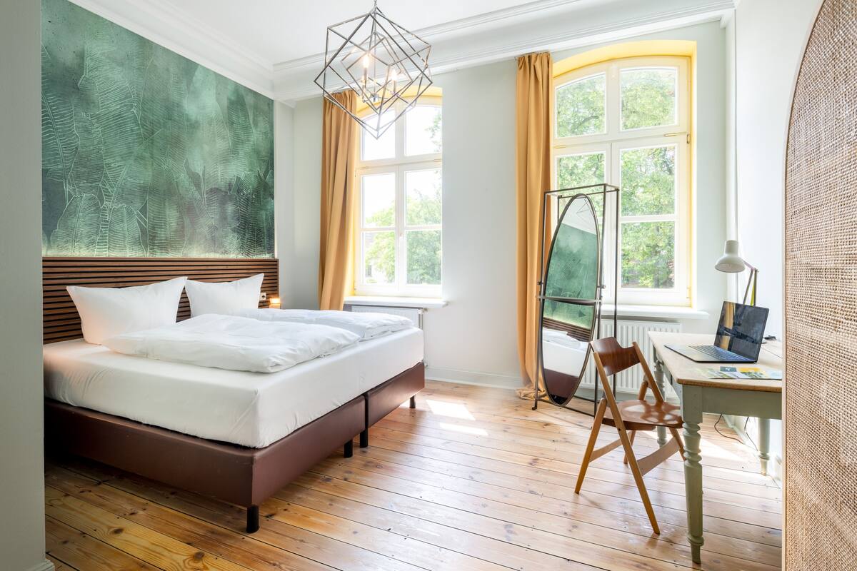 A modern bedroom with a large bed, a desk with a laptop, and a window with curtains.