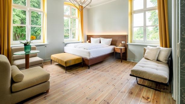 A bedroom with a large bed, a bedside table, a chair, and a sofa. There are windows with curtains and a view of greenery outside.