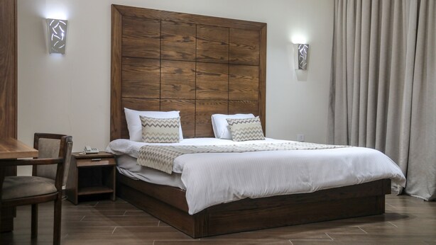 A bedroom with a wooden headboard, a bed with white bedding, a bedside table, a chair, and a curtain. - Eden Hotel (Jdeideh)