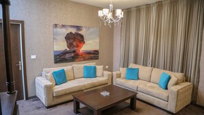 A living room with two beige sofas, a wooden coffee table, a chandelier, and a large painting of a volcanic eruption. - Eden Hotel (Jdeideh)