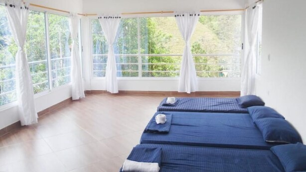 A room with three beds, large windows with white curtains, and a view of greenery outside.