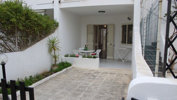 A courtyard with a white wall, a small garden, a table with chairs, and a door with shutters. - Trilocale Glicine piano terra Baia Verde (Gallipoli)
