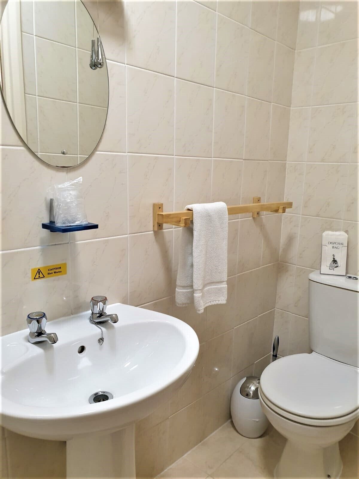 Et badeværelse med vask, spejl, håndklædeholder, toilet og et fareskilt for varmt vand.