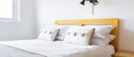 Una cama con ropa de cama blanca y almohadas decorativas, cabecera de madera, mesita de noche con lámpara y ventana con persianas.