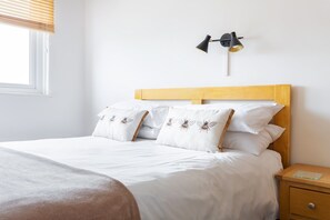 Una cama con ropa de cama blanca y almohadas decorativas, cabecera de madera, mesita de noche con lámpara y ventana con persianas.