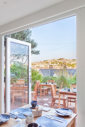 Un comedor con una mesa dispuesta para cuatro personas, vistas a un pueblo en la ladera a través de un ventanal, y cielo despejado.