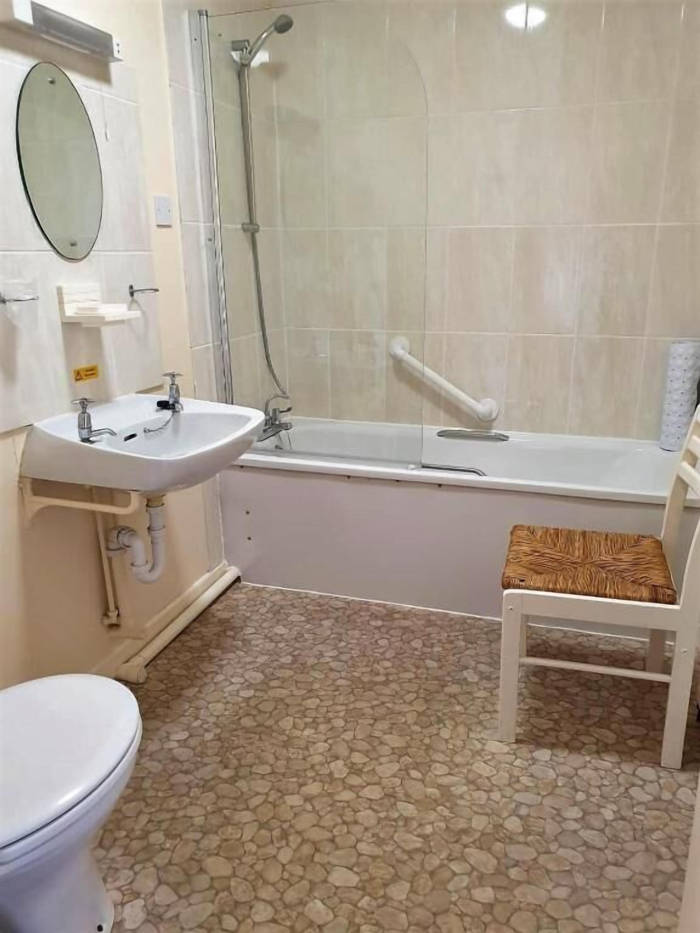 A bathroom with a toilet, sink, shower, and a chair.
