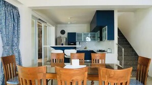 A dining area with a glass table and wooden chairs, a kitchen with dark cabinets, and a staircase leading to an upper level.