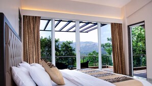 A bedroom with a large bed, a view of greenery and mountains, and a balcony with a chair.