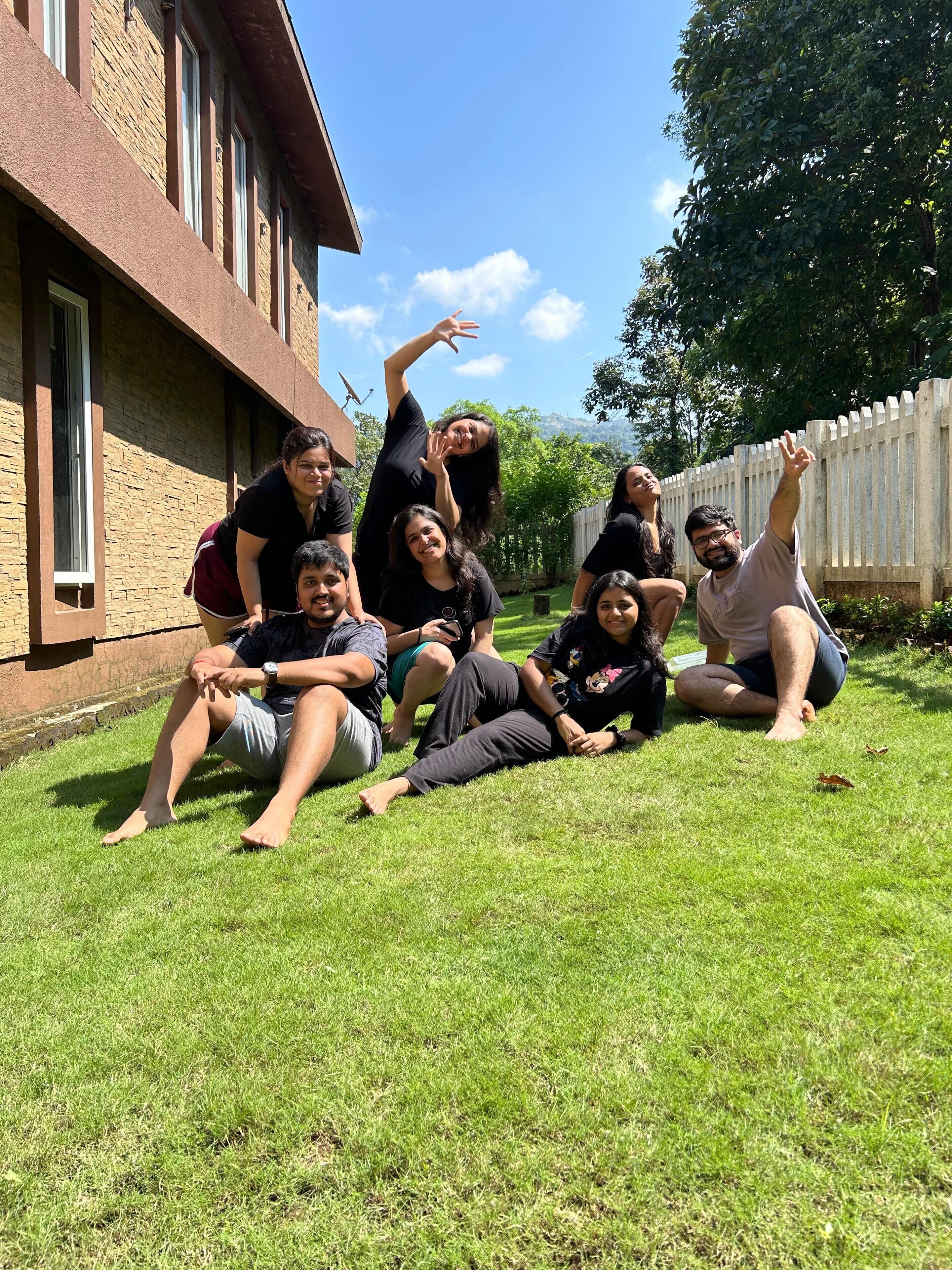Un groupe de personnes est assis sur l’herbe devant un bâtiment en briques, délimité par une clôture blanche.