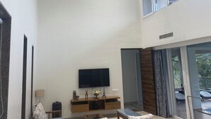 A spacious living area with a modern chandelier, a flat-screen TV, wooden furniture, and large windows offering a view of the outdoors.