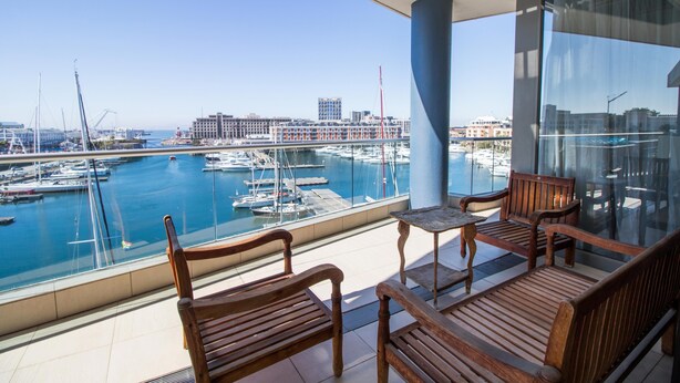 A balcony with wooden furniture overlooking a marina with boats and buildings in the background.