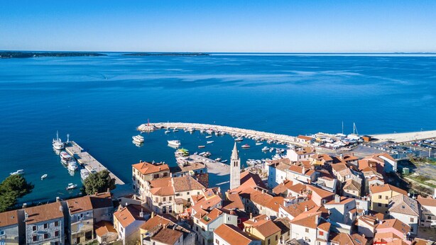 Vue aérienne d’une ville côtière avec un port de plaisance, des bâtiments aux toits recouverts de tuiles rouges et une église blanche imposante.