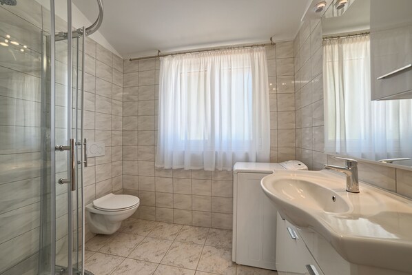 A bathroom with a glass shower enclosure, a toilet, a washing machine, and a sink with a mirror.
