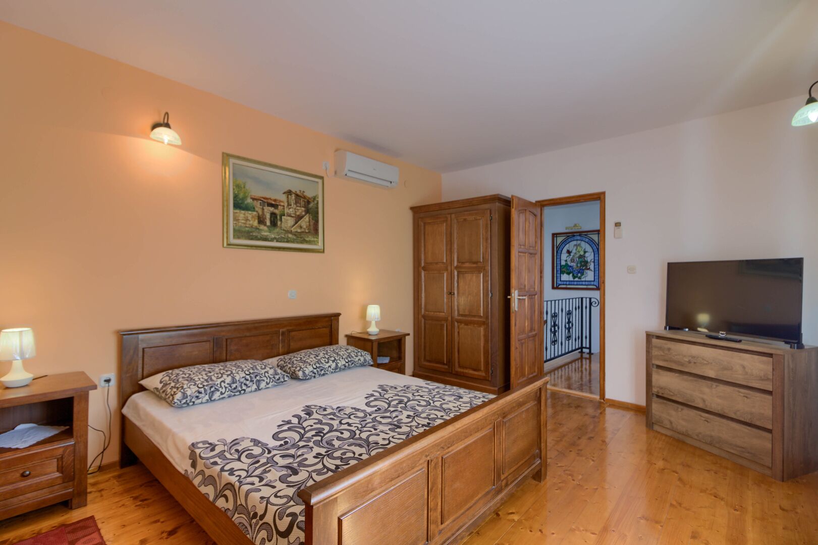 A bedroom with a bed, wooden wardrobe, TV stand, and a painting on the wall.
