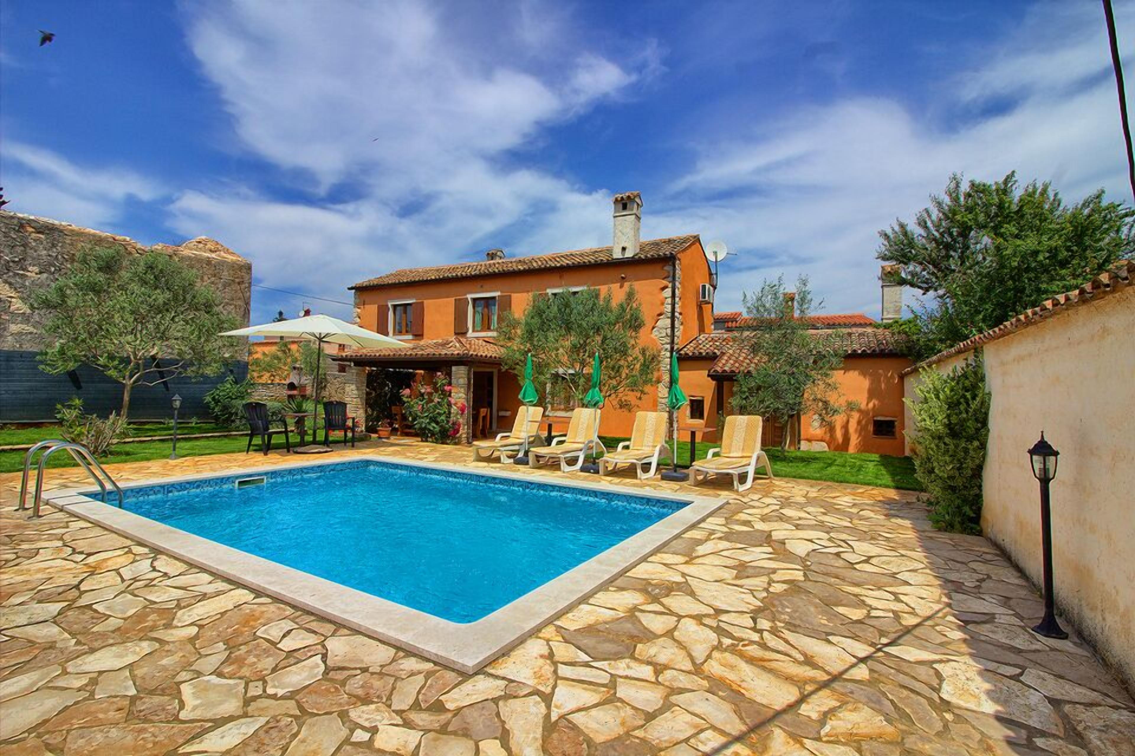 A swimming pool with stone tiles, a patio with lounge chairs, and a house with a tiled roof.