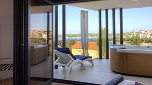 A balcony with a view of a coastal town, a hot tub, and a lounge chair.