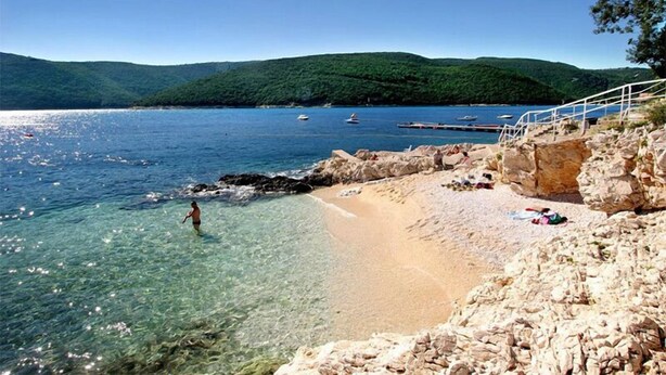 Ein Strand mit klarem Wasser, felsigem Ufer und Booten in der Ferne.