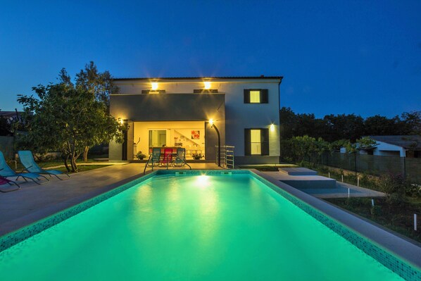 A modern house with a swimming pool, a covered patio, and a well-lit interior.