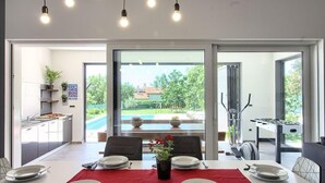 A dining area with a table set for four, a view of the outdoor pool and patio, and a kitchen area visible to the left. - Villa Medulin (Istria - Medulin) (Medulin)