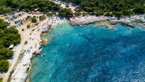Vista aérea de una zona costera con playa, aguas azul turquesa y un estacionamiento con coches.