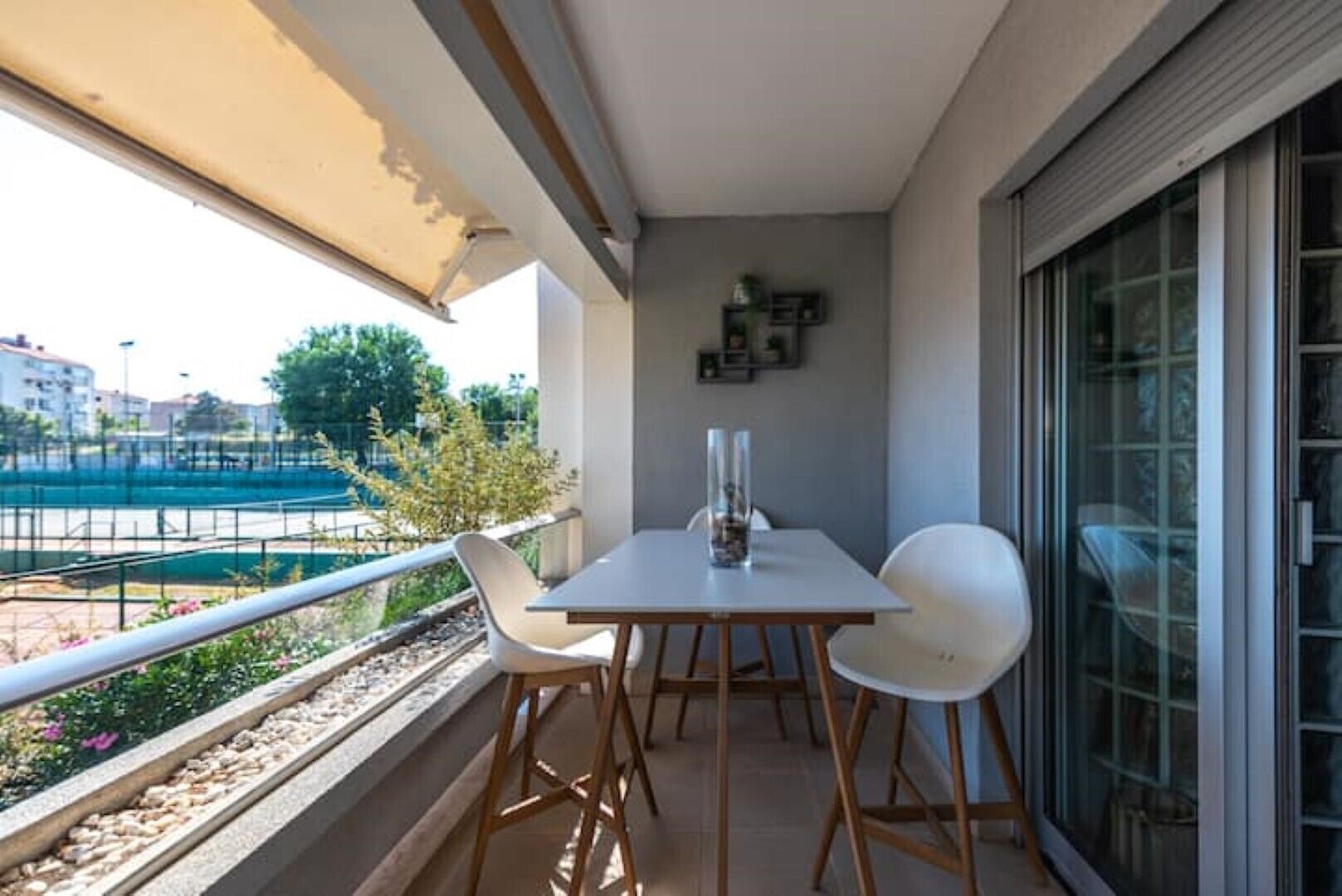 Un balcón con una mesa, sillas, un jarrón y vista a una cancha de tenis.