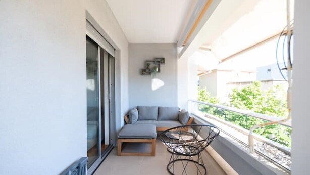 Un balcón con un sofá, una silla y una puerta corrediza de vidrio, con vista a zonas verdes.