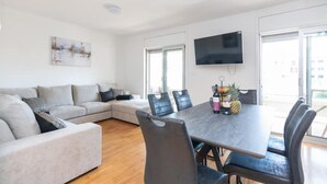 Una sala de estar moderna con un sofá gris, una mesa de comedor con sillas, un televisor de pantalla plana montado en la pared y un ventanal que proporciona luz natural.