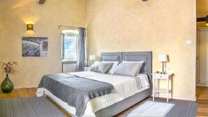 Ein Schlafzimmer mit einem großen Bett, zwei Nachttischen mit Lampen, einem Fenster mit Vorhängen, einem Gemälde an der Wand und einer Vase mit Trockenblumen.