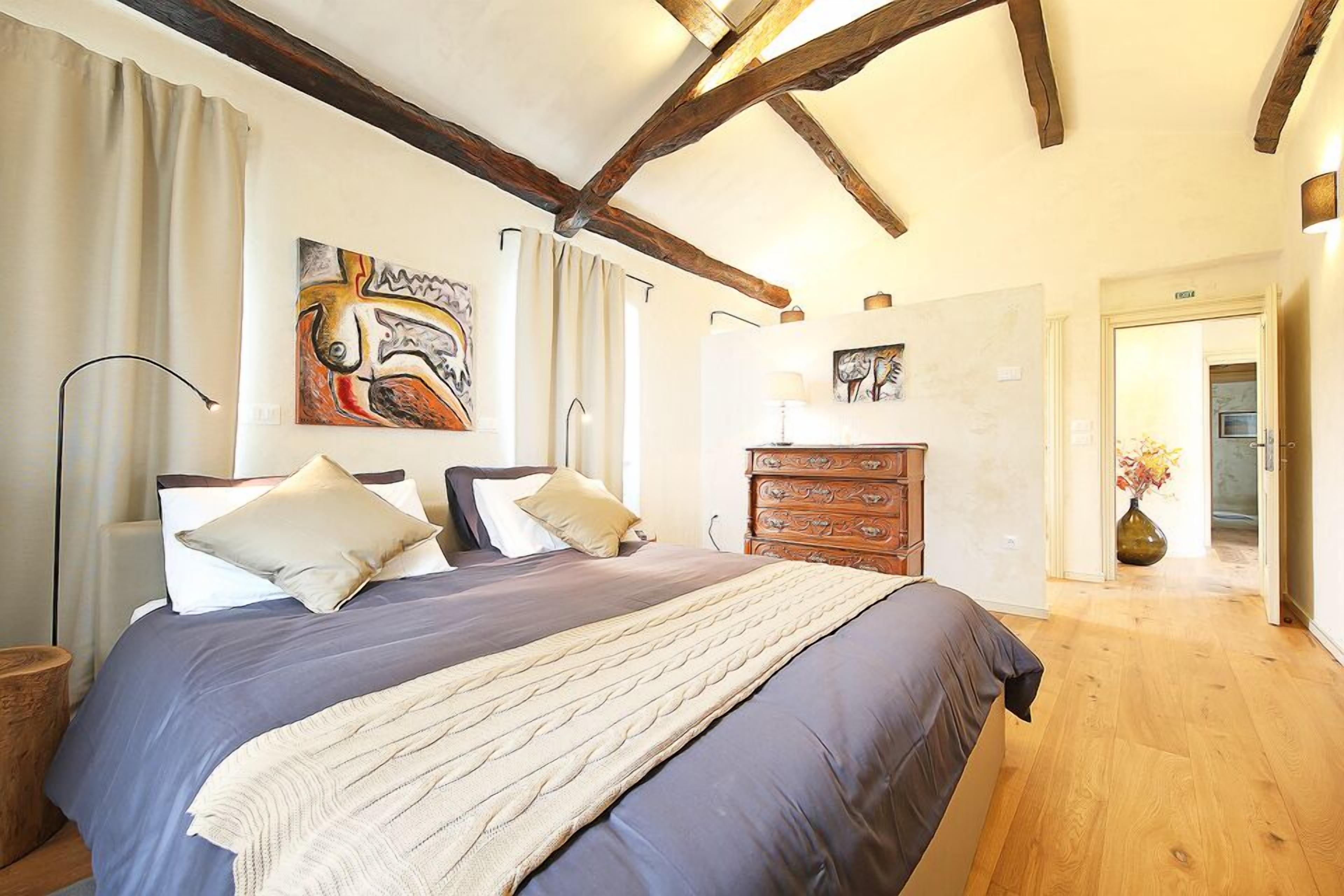 Un dormitorio con una cama grande, vigas de madera en el techo, un aparador y obras de arte en las paredes.
