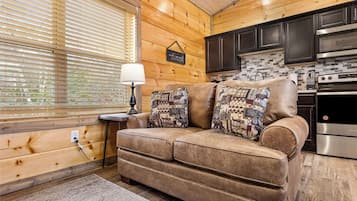 A cozy cabin interior with a brown leather sofa, a small side table with a lamp, and a kitchen area with dark cabinets and a stove.