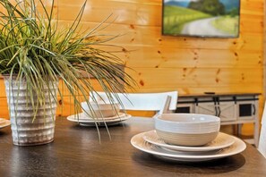Une table à manger en bois, avec des assiettes et des bols blancs, une plante en pot et un tableau accroché au mur.