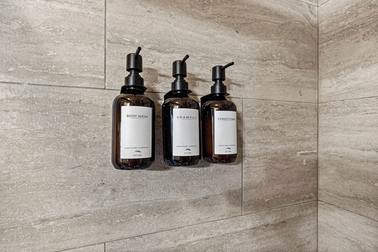 Three bottles of body wash, shampoo, and conditioner on a tiled wall.