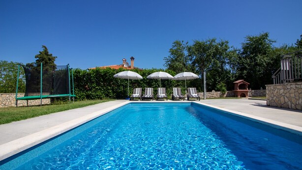 A rectangular swimming pool with clear blue water, surrounded by a stone wall and a grassy area. There are three lounge chairs with umbrellas and a small brick structure in the background. - Villa Marina (Istria - Paradiž) (Paradiž)