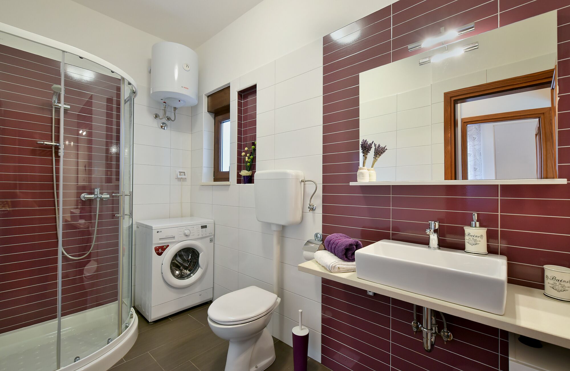 A modern bathroom with a glass shower enclosure, a washing machine, a toilet, and a vanity with a mirror.