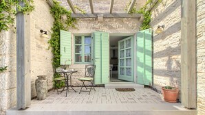 A stone building with a patio area featuring a table and chairs, a potted plant, and a door with green shutters.