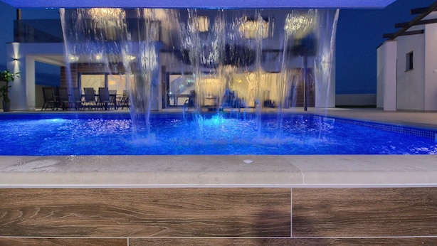 A swimming pool with a water feature in the center, surrounded by a wooden deck and a building with a glass facade. - Villa LUKA (Istria - Loborika) (Loborika)