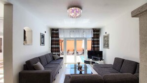 A modern living room with two sofas, a coffee table, and a TV. There is a large window with curtains and a view of the outside.