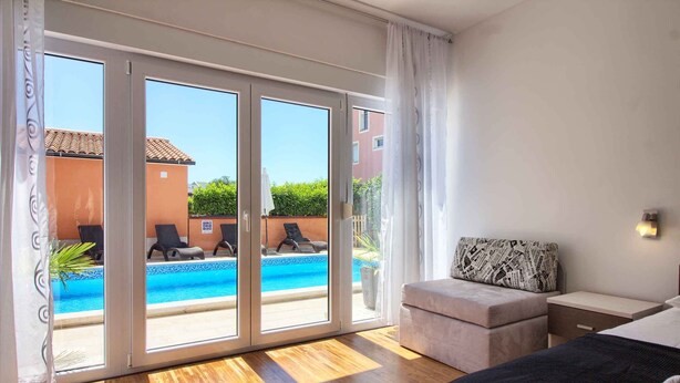 A hotel room with a large sliding glass door leading to a pool area, a bed, a bedside table, and a small seating area with a patterned pillow.