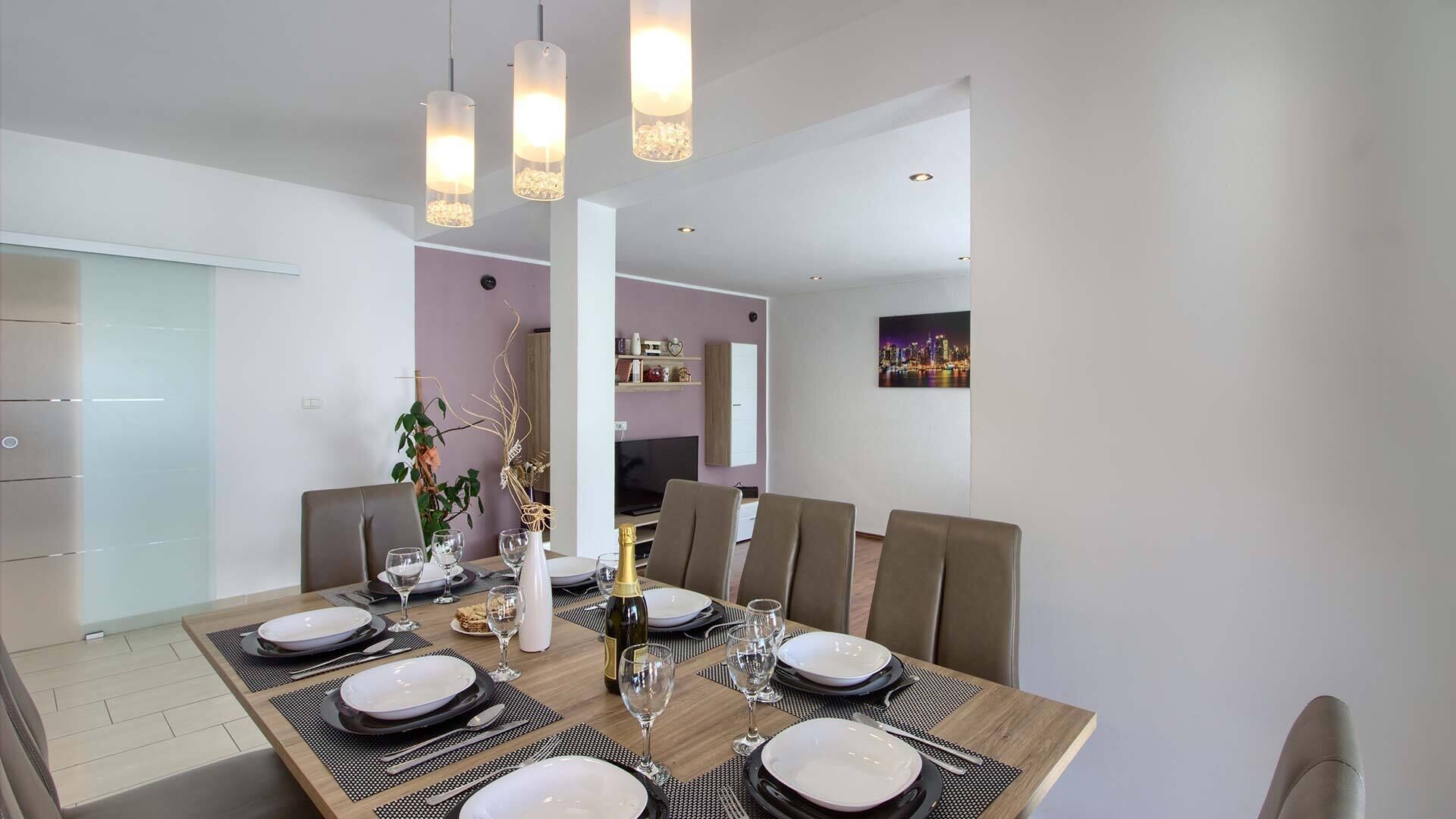 A modern dining area with a wooden table set for six, featuring black and white plates, glasses, and cutlery. There are three pendant lights above the table, and a cityscape painting on the wall.