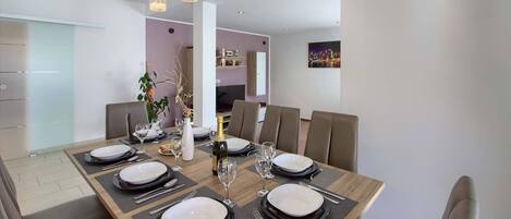 A modern dining area with a wooden table set for six, featuring black and white plates, glasses, and cutlery. There are three pendant lights above the table, and a cityscape painting on the wall.